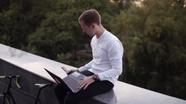 Genç adam gri parapetin üzerinde açık havada bacaklar dizüstü bilgisayar ile oturup, bir smartphone kullanarak konuşan beyaz gömlekli. Fırsat her yerde, serbest çalışmak zorunda. Arka planda yeşil ağaçlar