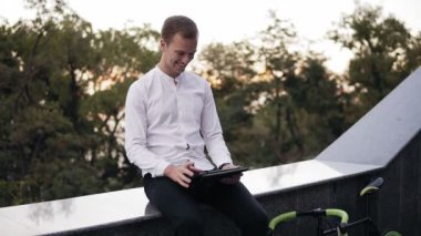Beyaz gömlek harcama zaman istimal tablet radyo sarışın genç adam. Merdivenlerden yakınındaki parapetin üzerinde oturan gülümseyen, mutlu adam. Arka planda yeşil ağaçlar