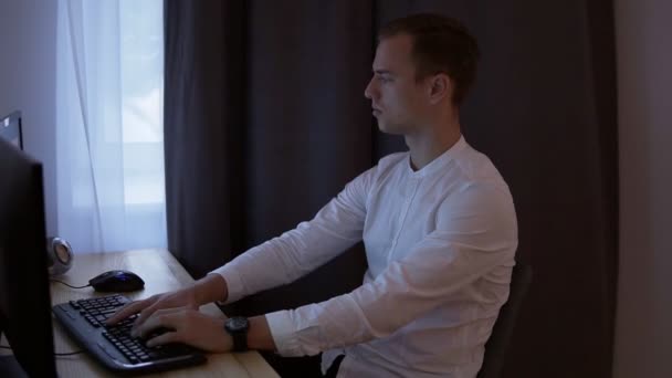 Hombre de negocios casual o frelancer que trabaja en casa, sentado en el escritorio, escribiendo en el teclado, mirando la pantalla de la computadora. Cortina gris en el fondo. Vista lateral — Vídeos de Stock