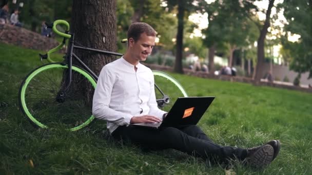 Ung framgångsrik affärsman i vit skjorta. Man sitter på gräset marken, arbete på bärbar pc dator i stadsparken på grön gräsmatta utomhus på naturen lutar på sin cykel snyggt trädet — Stockvideo