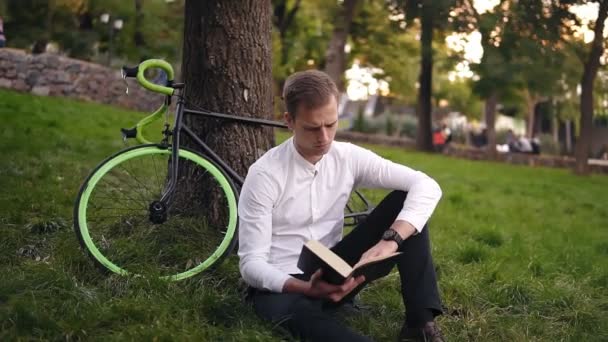 Joven hombre caucásico serio leyendo libro, sentado en el parque urbano de la ciudad de hierba, estudiando o leyendo, tiempo libre en el parque de la ciudad. Vista frontal — Vídeo de stock