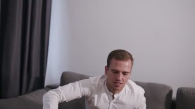 Beyaz gömlek ve siyah troussers beyaz delikanlı bacaklarına tutarak onun laptop üstünde belgili tanımlık gri kanepe evde ve başlangıç iş oturur. Arka plan üzerinde beyaz renkli duvar