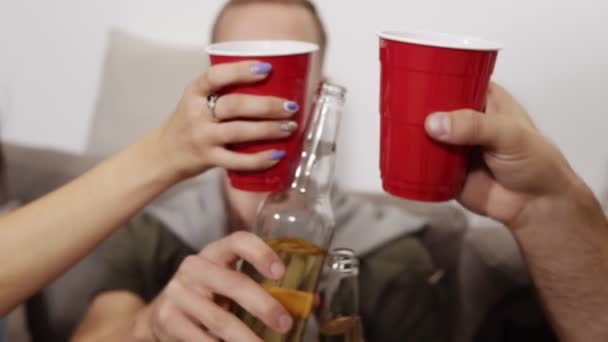 Dirigido a imágenes de cerca de vítores con botellas de bebida y tazas de plástico rojo. Los amigos se reúnen y se divierten en la sala de estar con el interior del loft, todo el mundo bebiendo cerveza o refresco — Vídeo de stock