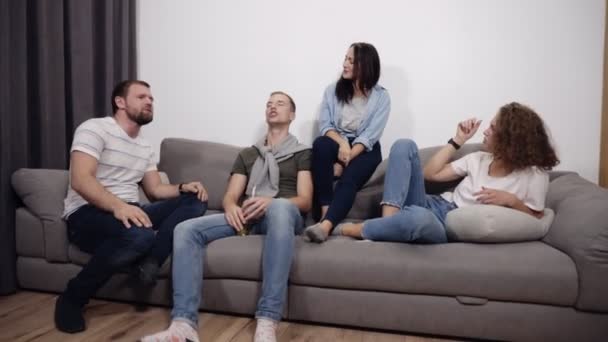 Чотири молоді смішні люди танцюють, сидячи на дивані. Друзі розважаються у вітальні в домашніх умовах концепції. Інтер'єр лофт — стокове відео