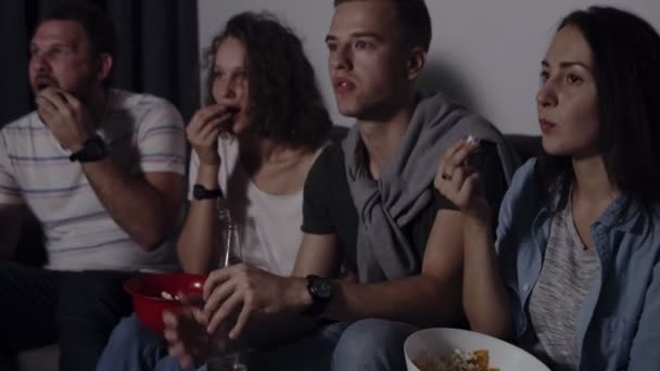 Vänner av fyra sit soffan, titta på rolig film på Tv. kaukasiska unga gruppen sitter på soffan, drinkar och snacks - greppa popcornen från röda skålen. Filmkväll — Stockvideo