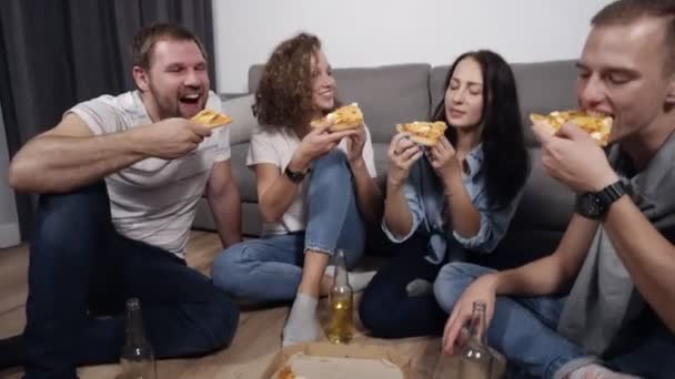 Grupo de quatro amigos se divertindo, pediu pizza grande e comer, rindo e falando, sentado no chão na sala de loft de cor cinza e branca. Beber e passar tempo juntos — Vídeo de Stock
