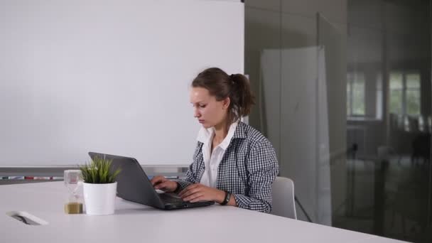 ポニーテールの少女は、モダンなロフト スタジオでラップトップを使用します。進行中の作業を研究しています。創造的なスタートアップで働く若いビジネス女性。小さな緑の花と後ろに砂時計、ノート パソコン — ストック動画