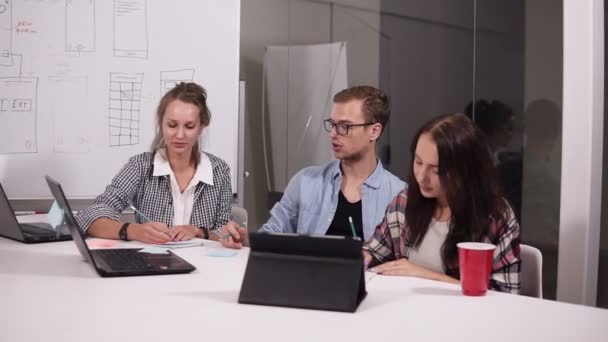 Business team discussing project or idea. Young man in glasses and two women in casual sitting at the office table with laptop and tablet on it in creative workplace — Stock Video