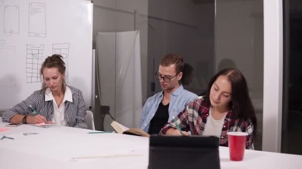 Tres empresarios o estudiantes sentados juntos trabajando en un escritorio de oficina comparando documentos, tomando notas. Joven en el medio leyendo un libro — Vídeo de stock