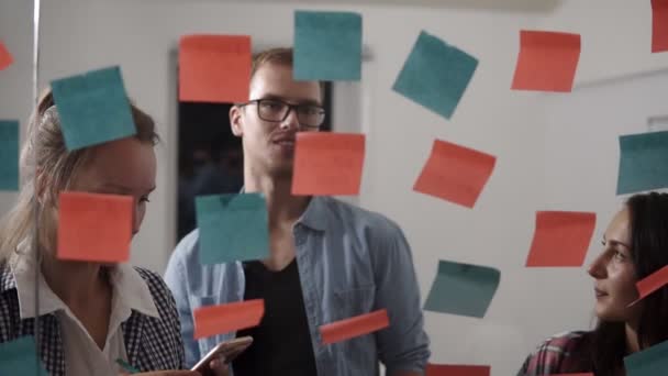 Rozmanitý tým pozitivní mladých lidí usmívající se přitom spolupracují během debaty a stojící za prosklenou stěnou s sticky barevné papíry. Veselí studenti učení slov od samolepky — Stock video