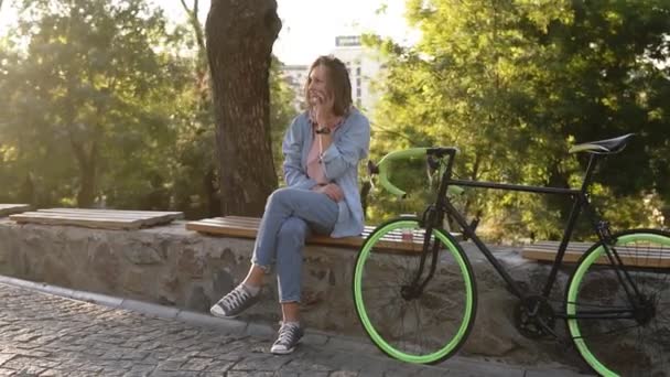 Linda chica sentada en el banco o parapeto en el parque de la ciudad con su bicicleta de trekking a su lado. Hablando por su teléfono móvil, sonriendo. Usar ropa casual de color rosa brillante y azul y zapatillas de deporte — Vídeo de stock