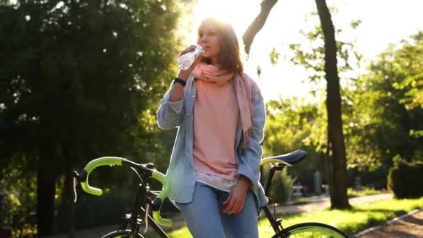 Menina bonita bebendo água clara de garrafa ao ar livre, apoiando-se em sua bicicleta de rodas de cor verde trekking. Fundo bonito da natureza, luz do sol cega a parte superior da imagem — Vídeo de Stock