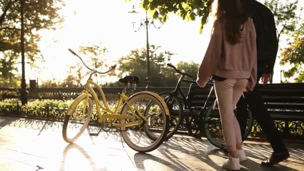 Casal jovem - homem em roupas casuais pretas e mulher de rosa vai andar de bicicleta no parque da cidade de verão. Vista rara — Vídeo de Stock