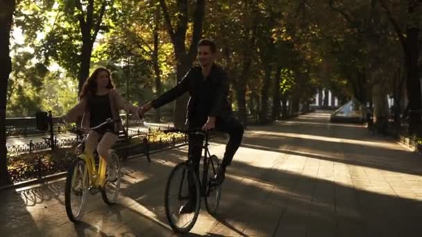 Vackra unga par ridning sina cyklar i tomma stadsparken eller boulevard sommartid. Håller händerna ihop medan Cykling cyklar. Framifrån — Stockvideo
