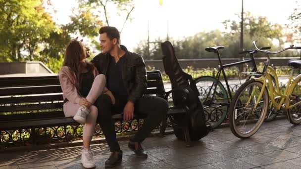 Chica bonita con el pelo castaño largo se sienta con su chico guapo morena en un banco del parque. Retrato de una pareja enamorada en un banco en el parque, mirándose con amor, tocándose. Caja de guitarra negra — Vídeo de stock