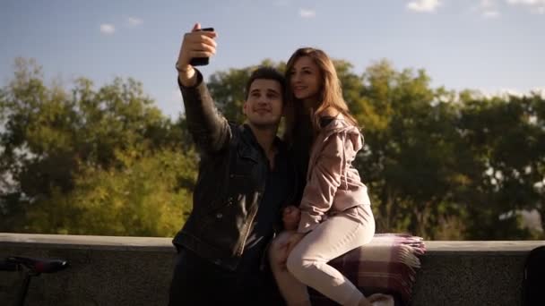 Lyckliga unga paret tar selfie av mobiltelefon på gatan medan flicka sitter på räcket. Vackra par selfie, kärlek och roligt koncept. Blå himmel och toppen av träden i bakgrunden — Stockvideo