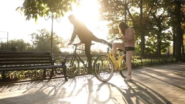 Hermosa pareja joven montando sus bicicletas en el parque o bulevar vacío de la ciudad en verano. Detente y siéntate en el banco y abrázate. Una pareja amorosa. El sol brilla en el fondo — Vídeo de stock