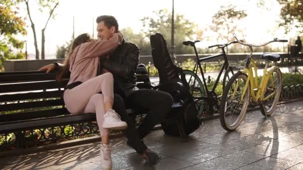 En vacker kaukasiska par sit embracing på en parkbänk. Porträtt av ett par i kärlek på en bänk i parken och att ha roligt tillsammans. Svart gitarrfodral och cykel bredvid dem. — Stockvideo