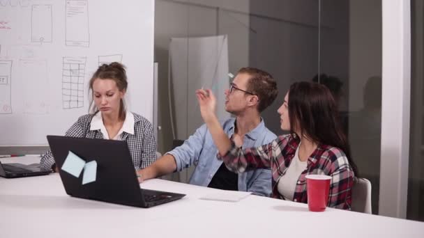 A fiatalember a szemüveg és a két nő egymással valamit megvitatni kreatív munkahelyi office asztalnál ülve. Barna nő mutató whitesheet tábla, látása megosztása — Stock videók
