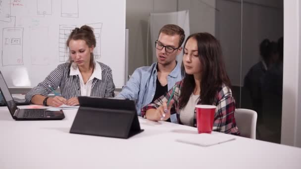 Молодой человек в очках и две женщины в повседневной жизни сидят за рабочим столом с ноутбуком и планшетом на нем в креативном рабочем месте. Человек посередине показывает карандашом на доске, объясняя свою — стоковое видео
