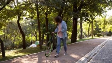 Önden görünümü dışında bir bisiklet sabah parkta yürüyen bir genç neşeli kadın. Bir dümen ile tutarak trekking bisikleti ile yürüyen kız. Ağaçlar ve güneş parlar arka plan üzerinde. Mercek parlaması