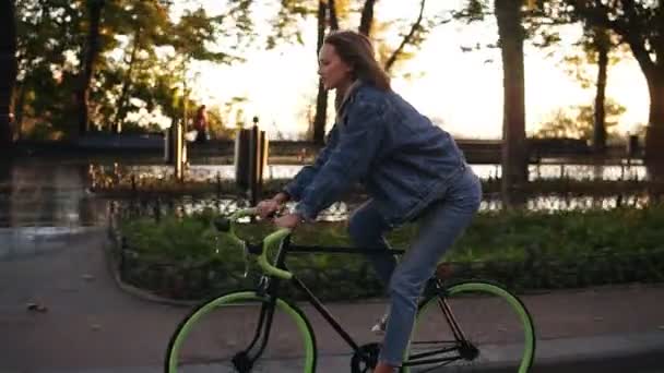 Menina atraente andando de bicicleta no parque da manhã ou avenida. Vista lateral de uma jovem mulher em roupas casuais montando uma bicicleta de trekking — Vídeo de Stock