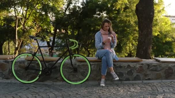 Задумчивая девушка, сидящая на скамейке в городском парке с велосипедом рядом с ней. Смотрю на ее мобильный, в яркой повседневной одежде. Деревья и солнце на заднем плане — стоковое видео