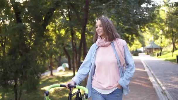 Ragazza felice e sorridente che cammina accanto a una bicicletta nel parco del mattino. La donna che cammina con la sua bici da trekking, reggendo con un timone. Alberi e sole splendono sullo sfondo. I brillamenti delle lenti. Indossare rosa — Video Stock