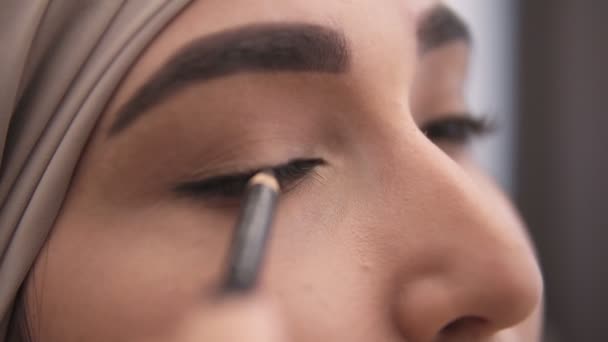 Vista extremadamente cercana de una joven musulmana haciendo una flecha en su párpado usando lápiz de ojos negro. Impresionante look de mujer musulmana en pañuelo beige. Vista lateral — Vídeos de Stock