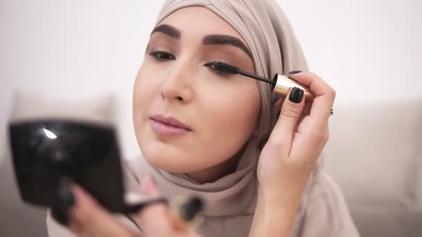 Atraktivní Muslimská žena dělá make-up profesionálně. Kartáč na řasy s řasenky. Na sobě béžový šátek. Bílá zeď na pozadí — Stock video