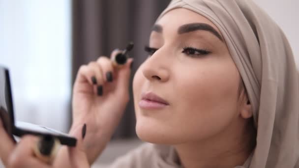 Hermosa mujer musulmana haciendo maquillaje profesionalmente. Cepille las pestañas con rímel. Usando pañuelo beige. Pared blanca en el fondo. Vista lateral — Vídeos de Stock