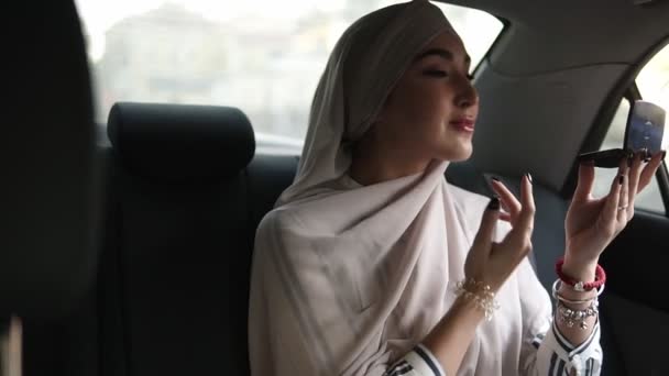 Retrato de uma jovem mulher muçulmana em lenço de cabeça bege, sentado no carro enquanto olha para um pequeno espelho cosmético e verificar sua maquiagem — Vídeo de Stock