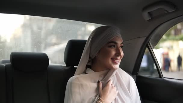 窓から外を見て、笑みを浮かべてし、カメラにポーズしながら車の中で座っているベージュのスカーフの若いイスラム教徒の女性の肖像画 — ストック動画