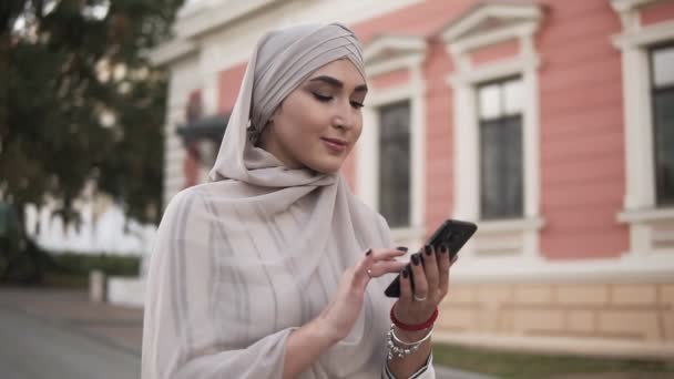 Muslim wanita cantik di hijab pastel dengan makeup berdiri di jalan memegang smartphone sementara berjalan-jalan di kota. Mengirim pesan — Stok Video