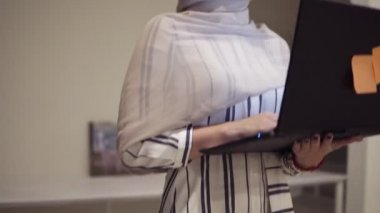 Siyah laptopunu elinde tutarken hall tarafından gidiş bir hijab giyen genç Arapça kadın. Çalışan, çalışma yeri, konferans salonu, koridor. Düşük açılı genel bakış görüntüleri