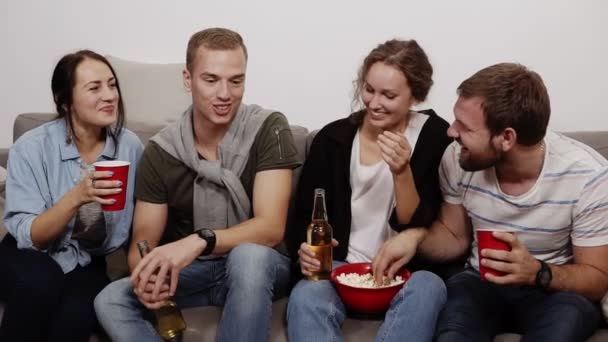 Jovens amigos alegres do sexo feminino e masculino com bebidas estão sentados no sofá em casa, comendo pipocas da tigela vermelha, fazendo piadas, rindo. Vista frontal — Vídeo de Stock