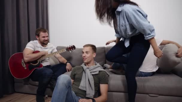 Accelererad bilder av vänner att ha roligt tillsammans i vardagsrummet. Skäggiga mannen sitter i soffan med akustisk gitarr, flickor sitter på soffan och en ung man på golvet med en flaska — Stockvideo