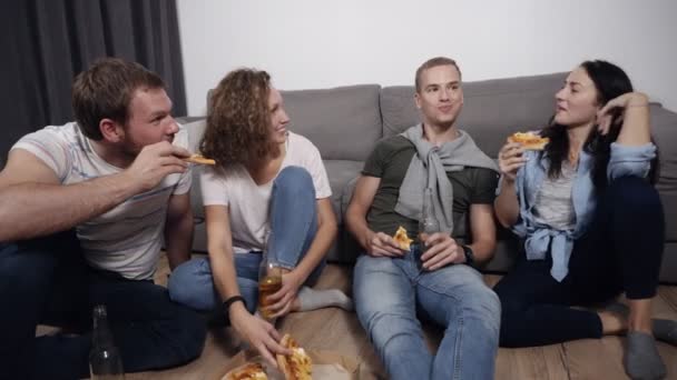 Concetto di cibo, svago e felicità: quattro giovani sorridenti che mangiano pizza a casa e bevono una birra seduti sul pavimento. Vista frontale — Video Stock