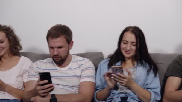 Diversos jóvenes sonriendo personas sentadas en fila en el sofá gris juntos obsesionados con los dispositivos en línea, adictos caucásicos utilizando sus teléfonos inteligentes. Fondo de pared blanco. Vista frontal — Vídeo de stock