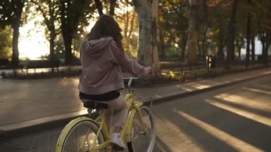 Sevimli küçük kız sürme sarı Bisiklet yaz şehir park Caddesi üzerinde renkli. Ağaçlar ve arka planda parlıyor. Görünümünde nadir