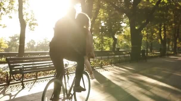 Αγαπώντας νεαρό ζευγάρι έχει τη διασκέδαση ιππασία στο ίδιο ποδήλατο σε υπαίθρια δραστηριότητα με ήλιο φωτισμό στο φόντο. Κορίτσι είναι κάθεται στο πηδάλιο ενώ ο φίλος της ποδηλασίας. Σπάνια θέα — Αρχείο Βίντεο