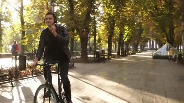 Kaukasische jongeman met koptelefoon fiets in het stadspark op de trekking zwart fiets. Paardrijden en een goed gevoel terwijl u luistert naar de favoriete muziek. Zon schijnt op de achtergrond — Stockvideo
