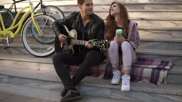 Joven pareja caucásica sentada en las escaleras y el hombre tocando la guitarra mientras está sentado con su novia al aire libre en las escaleras de la ciudad en el cuadros. Bicicletas a su lado. Luz solar — Vídeo de stock