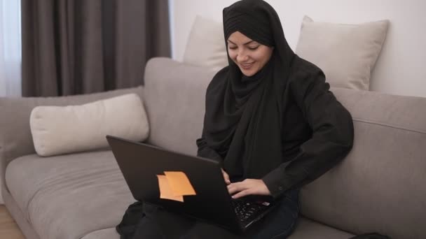 Joven mujer musulmana sonriente con ropa negra sentada en un sofá gris en casa y trabajando con su computadora portátil de rodillas, escribiendo, trabajando por cuenta propia en casa — Vídeo de stock