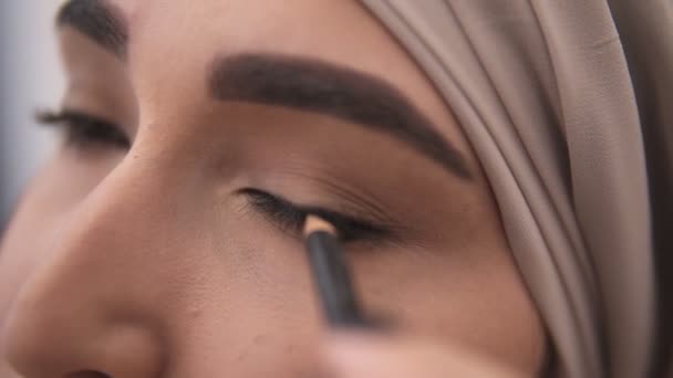 Visão extremamente próxima de uma jovem mulher muçulmana fazendo uma flecha na pálpebra usando um lápis de olho preto. Olhar deslumbrante de mulher muçulmana em lenço de cabeça bege — Vídeo de Stock