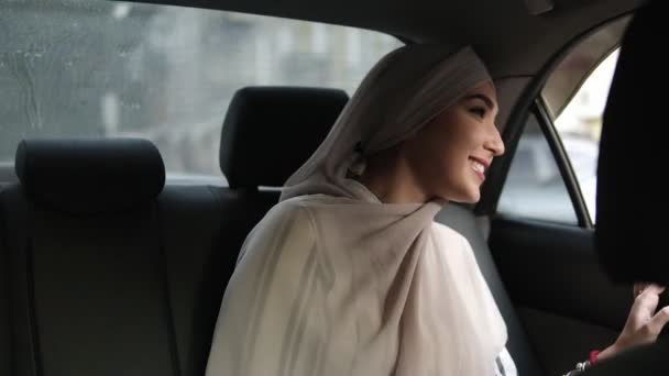 Retrato de una joven musulmana en pañuelo beige, sentada en el coche mientras mira por la ventana y sonríe — Vídeos de Stock