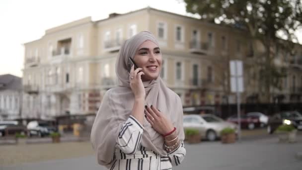 Mujer musulmana en hiyab con maquillaje caminando por la calle hablando por teléfono en un fondo de hermoso edificio clásico antiguo. La mujer está vestida con una ropa elegante, sonriendo. Vista lateral — Vídeo de stock