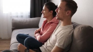 Genç çift kanepede gri evde parlak oturma odasında Tv izlerken. Heyecan verici kaç televizyona baktılar göstermek. Yan görünüm