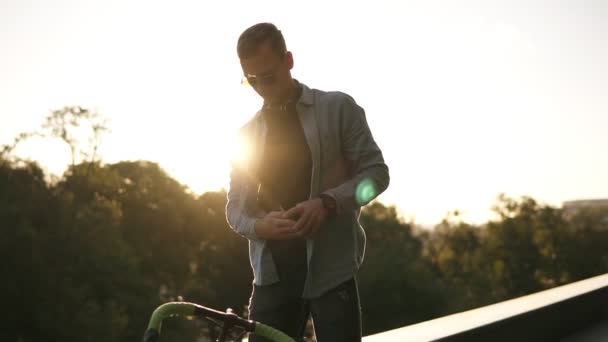 Ritratto di un giovane che sceglie la traccia musicale sul suo smartphone, indossa le cuffie e inizia a pedalare sulla sua classica bici vicino al parco cittadino. Sole forte splendente sotto gli alberi sullo sfondo — Video Stock