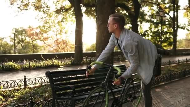 Fiatal férfi sötét farmer, kék inget és napszemüveg lovaglás saját kerékpár a városi Park egyedül. Megálló közelében a padon, és megteszi a zsebében a mobiltelefon — Stock videók